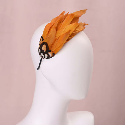 A gold yellow feather fascinator headpiece with brown, black and gold patterned feathers displayed on a white mannequin head against a soft pink background.