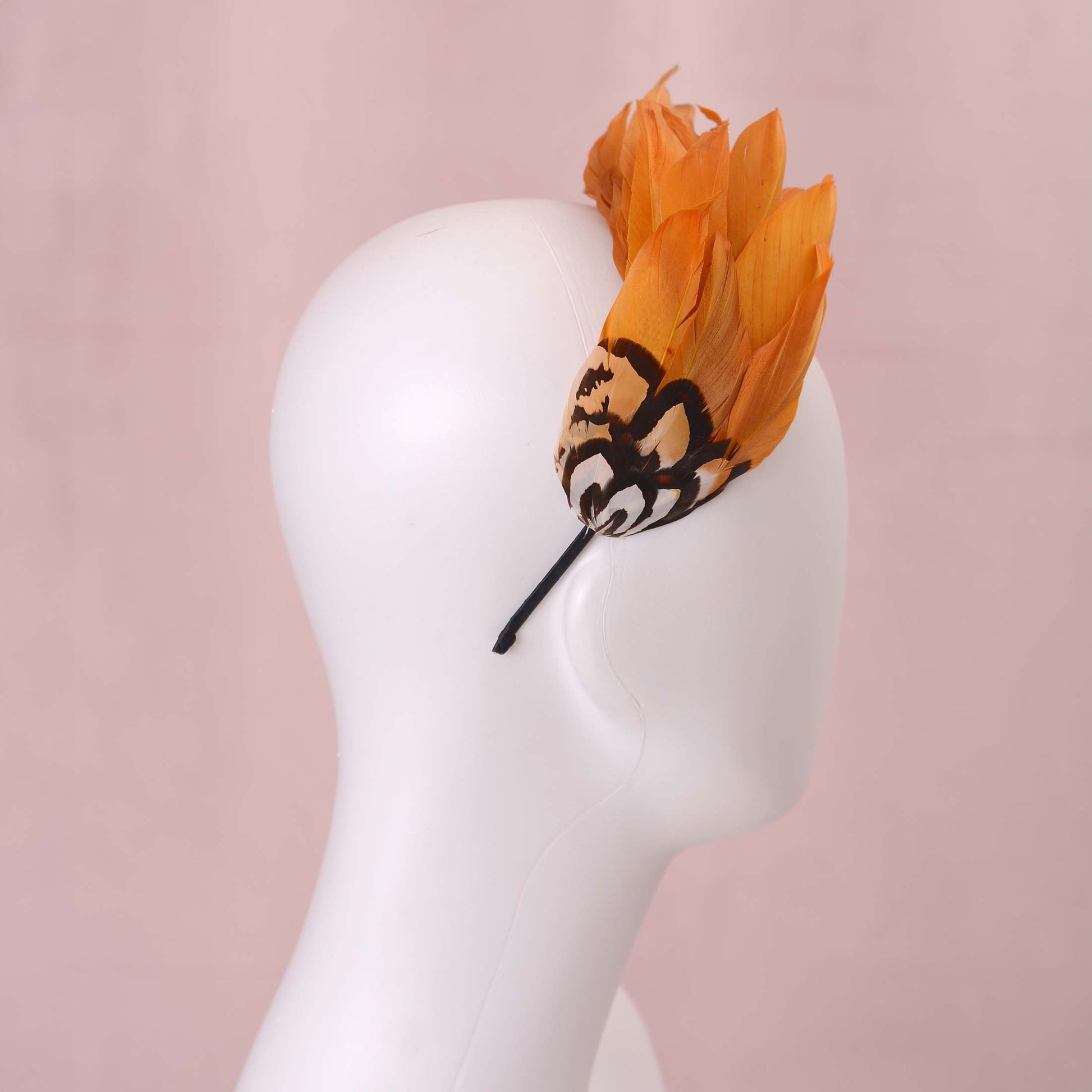 A gold yellow feather fascinator headpiece with brown, black and gold patterned feathers displayed on a white mannequin head against a soft pink background.