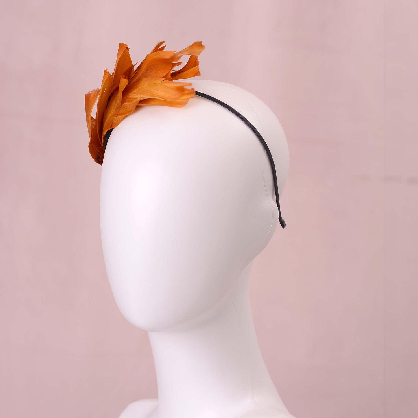 A gold yellow feather fascinator headpiece with brown, black and gold patterned feathers displayed on a white mannequin head against a soft pink background.