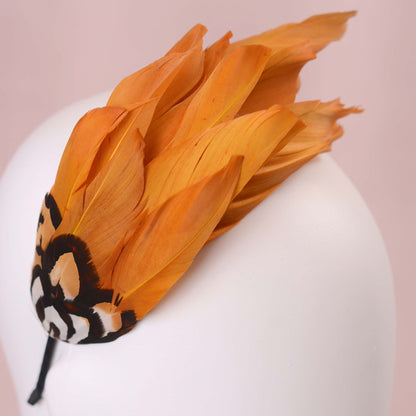 A  close up of a gold yellow feather fascinator headpiece with brown, black and gold patterned feathers displayed on a white mannequin head against a soft pink background.