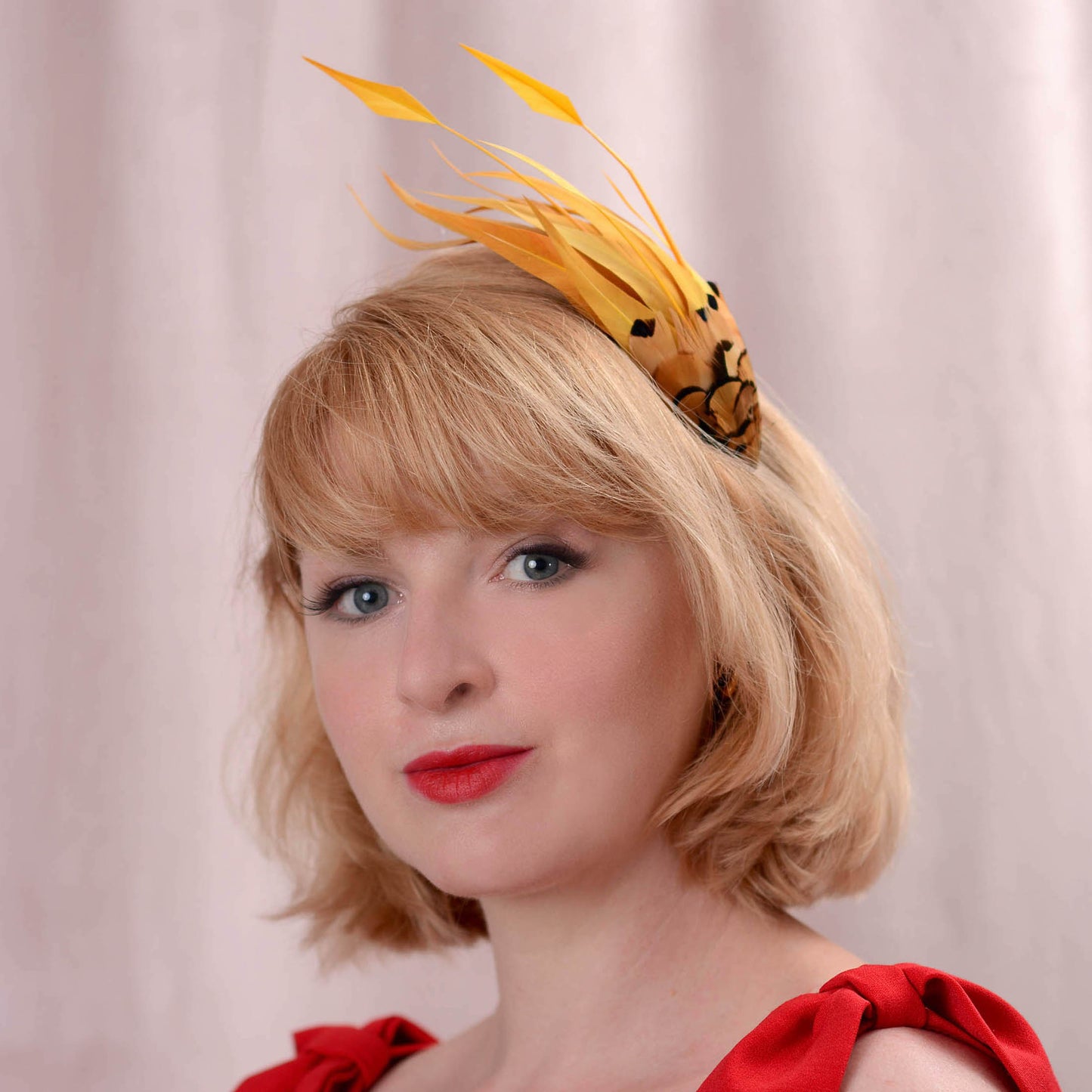 A blonde woman with a short bob hairstyle wearing a yellow and black feather fascinator headpiece, paired with a red top, set against a soft pink curtain background.