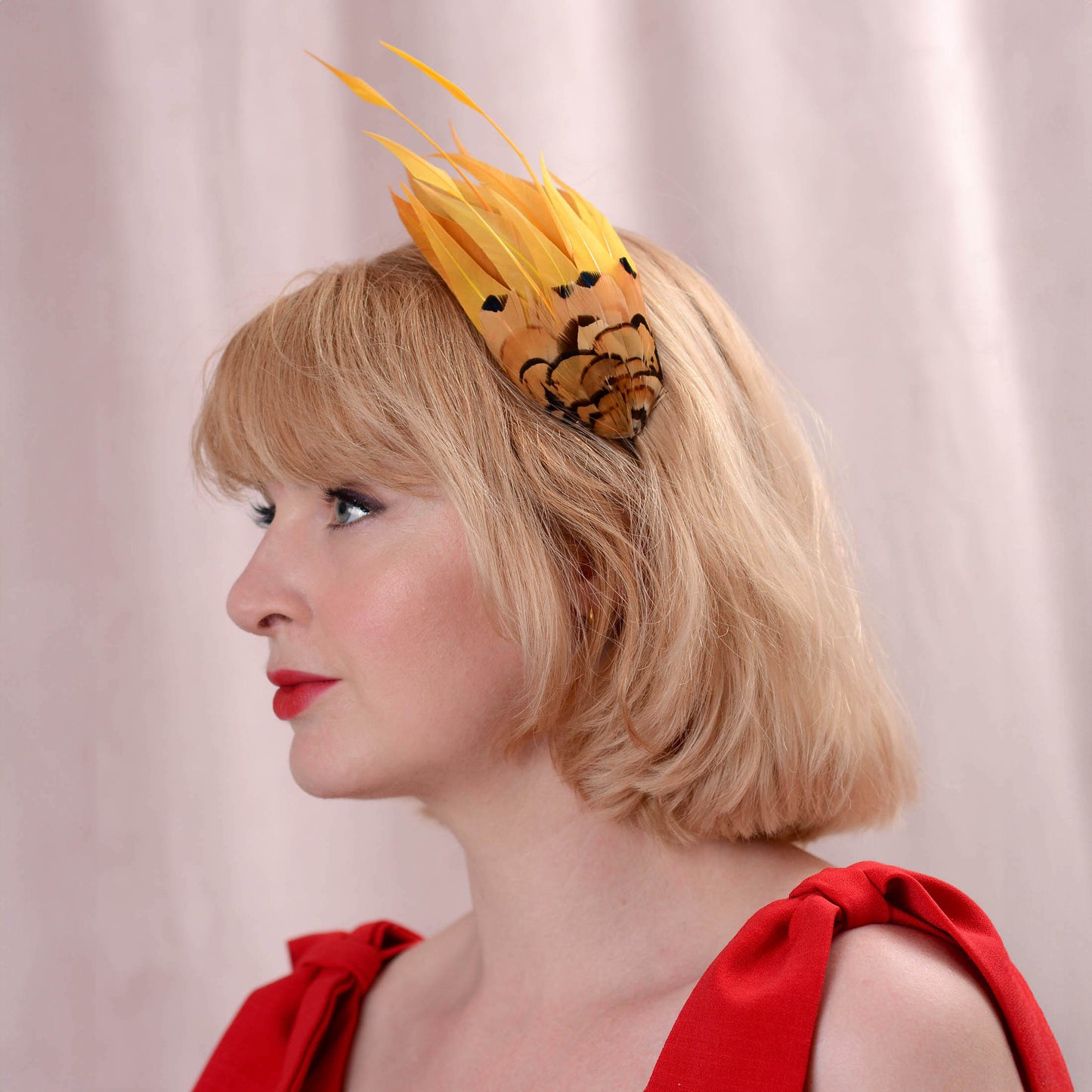 A woman in profile wearing a gold yellow fascinator headpiece with black and gold floral detailing, paired with a red dress against a soft pink curtain background.