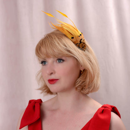 A woman with blonde hair wearing a gold yellow feather fascinator headpiece and a red dress, photographed against a soft beige fabric backdrop.
