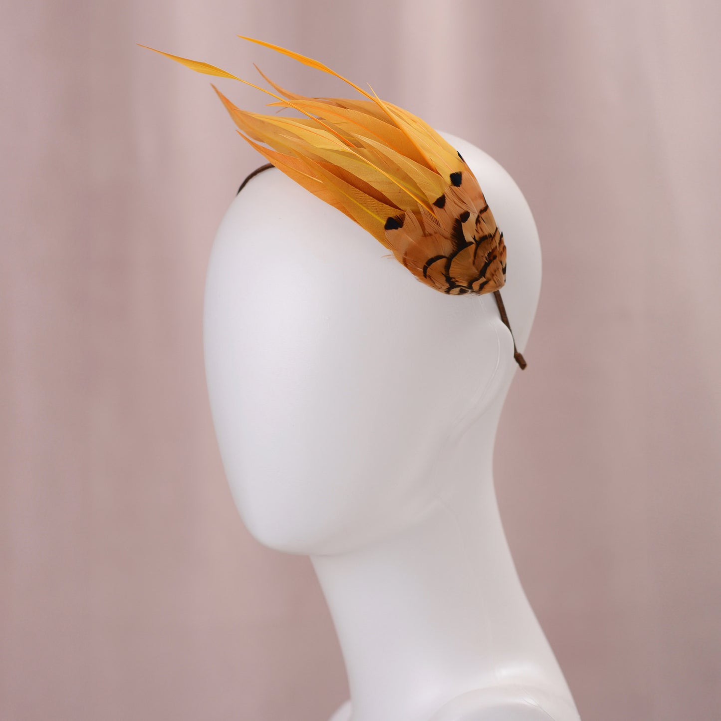 A decorative fascinator headpiece with long gold yellow feathers and a patterned brown and gold yellow base, displayed on a white mannequin head against a soft pink background.