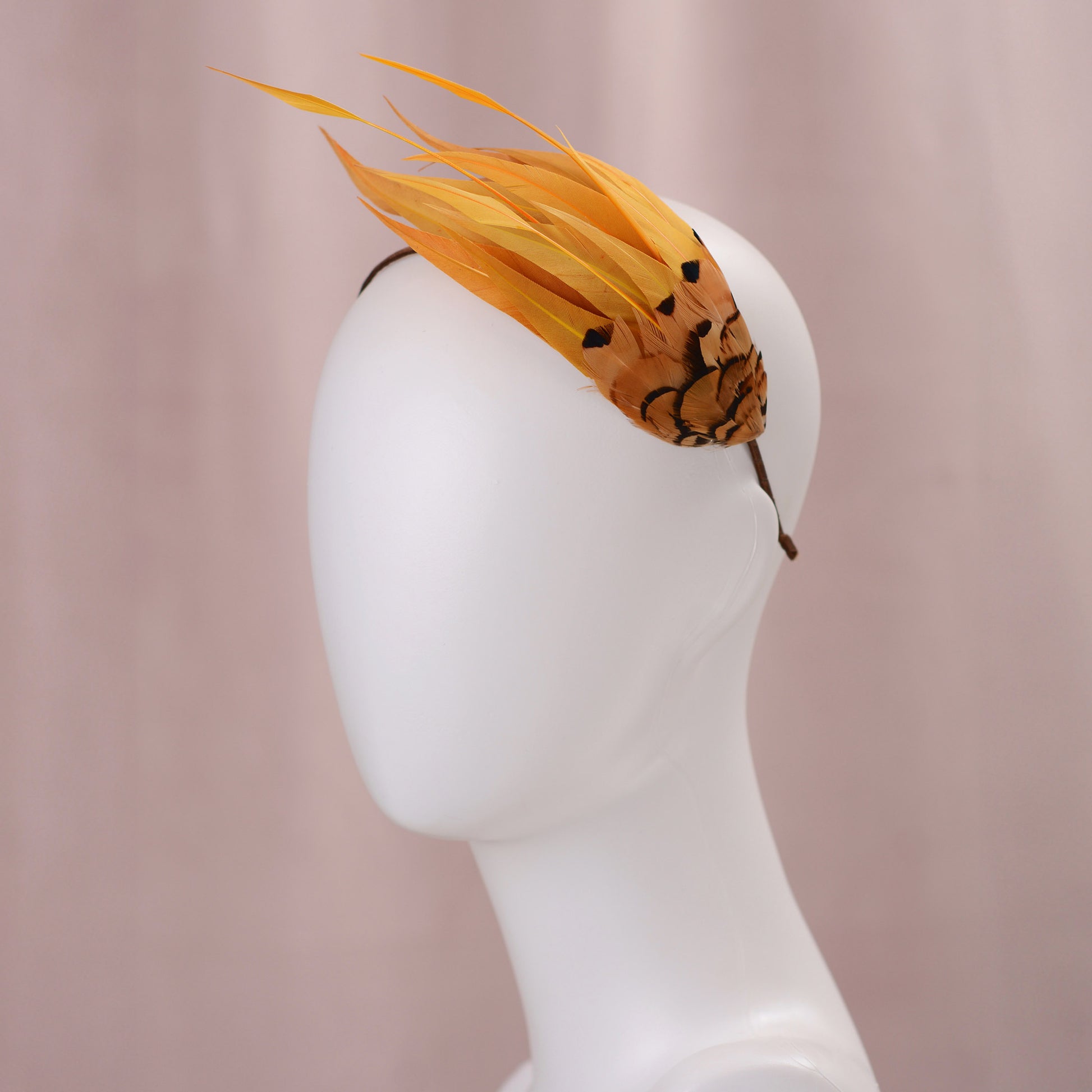A decorative fascinator headpiece with long gold yellow feathers and a patterned brown and gold yellow base, displayed on a white mannequin head against a soft pink background.