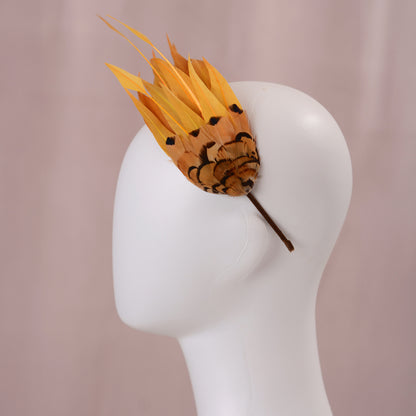 A decorative feather fascinator with bright golden yellow feathers and a brown patterned base, displayed on a white mannequin head against a soft pink background.