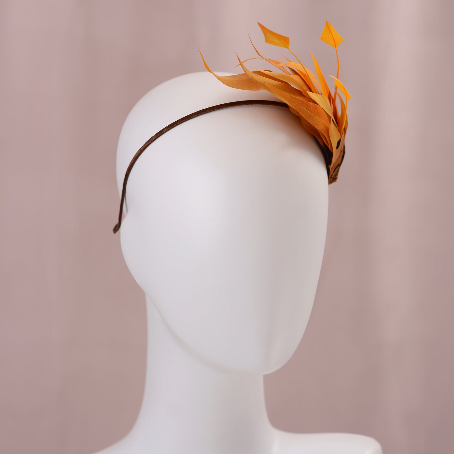 A brown headband with a decorative side perching arrangement in gold-yellow feathers, displayed on a white mannequin head against a soft pink background.