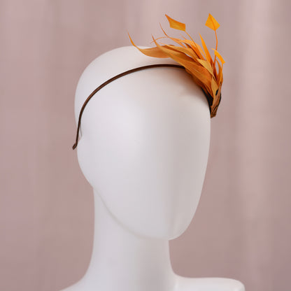 A brown headband with a decorative side perching arrangement in gold-yellow feathers, displayed on a white mannequin head against a soft pink background.