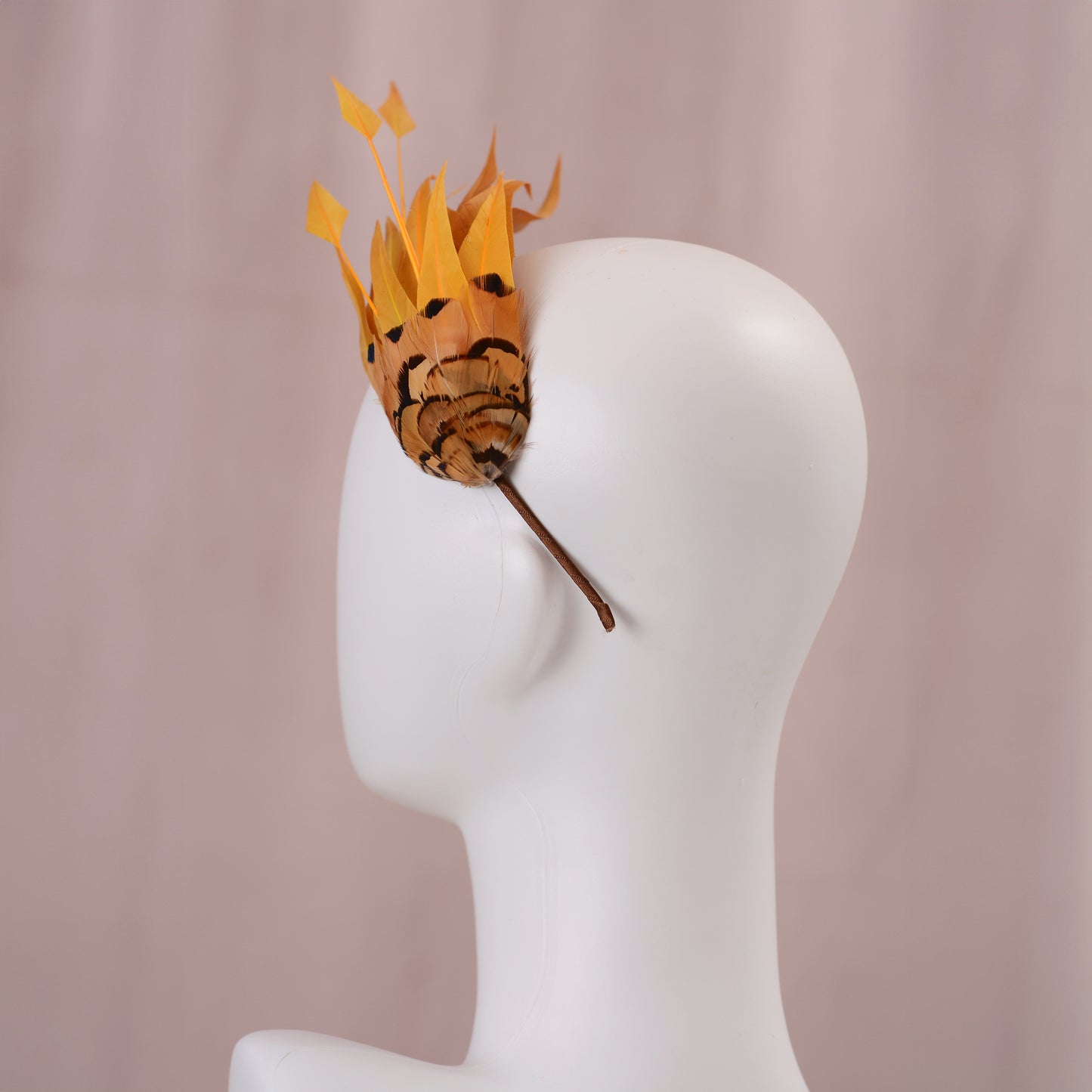 A decorative headpiece made from gold  yellow feathers, displayed on a white mannequin head against a soft pink background.