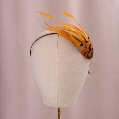 A decorative fascinator headpiece with long saffron yellow feathers and a patterned brown and black finial, displayed on a cream fabric mannequin head against a soft pink background.
