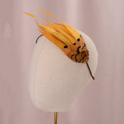A decorative feather fascinator headpiece in yellow and orange-brown with black pattern details, displayed on a cream fabric mannequin head against a soft pink background.