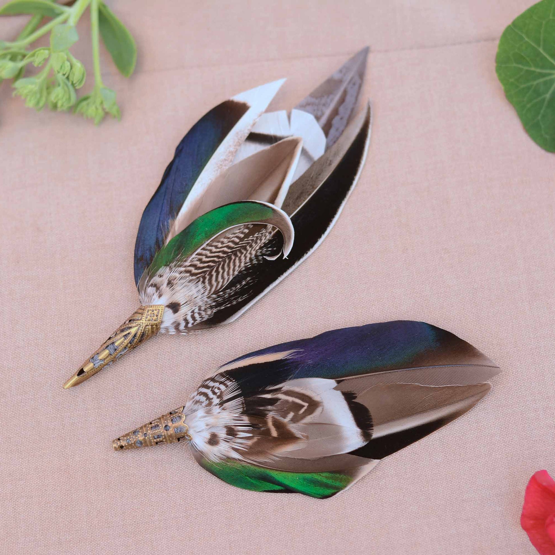 Pair of decorative feather wedding boutonnieres  in complimenting styes shown on a beige fabric background, labeled Style 1, Style 2, and Style 3. The feathers have iridescent blue-green and green sections, brown and white patterned shafts, and metallic gold tips.