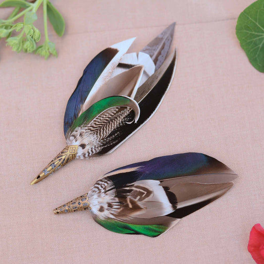 Pair of decorative feather wedding boutonnieres  in complimenting styes shown on a beige fabric background, labeled Style 1, Style 2, and Style 3. The feathers have iridescent blue-green and green sections, brown and white patterned shafts, and metallic gold tips.