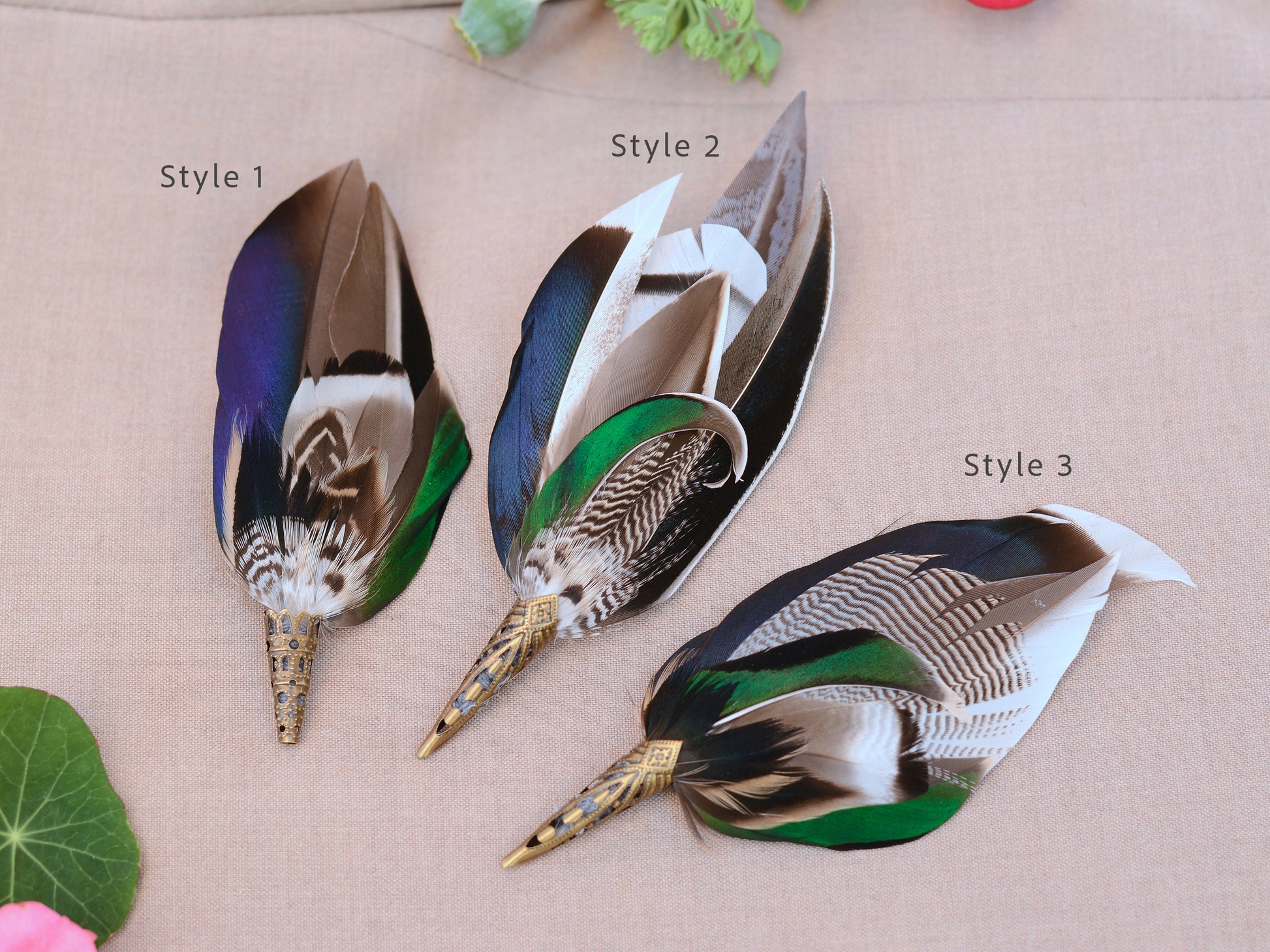 Three decorative feather wedding boutonnieres  in complimenting styes shown on a beige fabric background, labeled Style 1, Style 2, and Style 3. The feathers have iridescent blue-green and green sections, brown and white patterned shafts, and metallic gold tips.