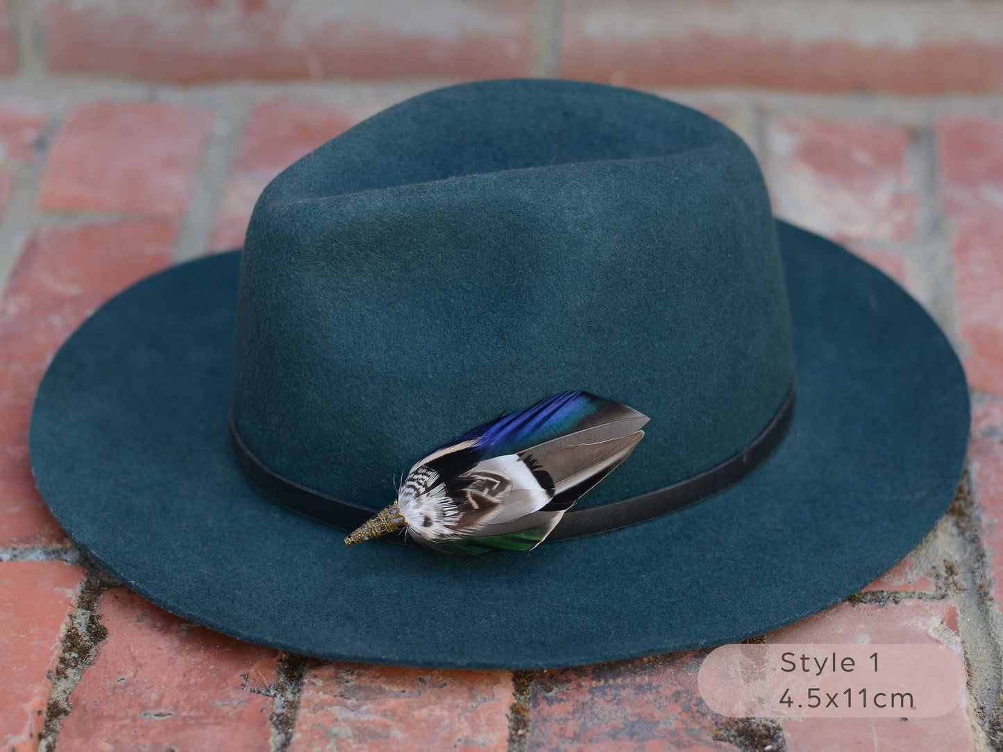 Duck feather hat pin with iridescent blue and green feathers on a dark teal hat with a thin black leather hat band on a red brick surface.