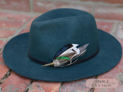 Large duck feather hat pin with iridescent blue and green feathers on a dark teal hat with a thin black leather hat band on a red brick surface.