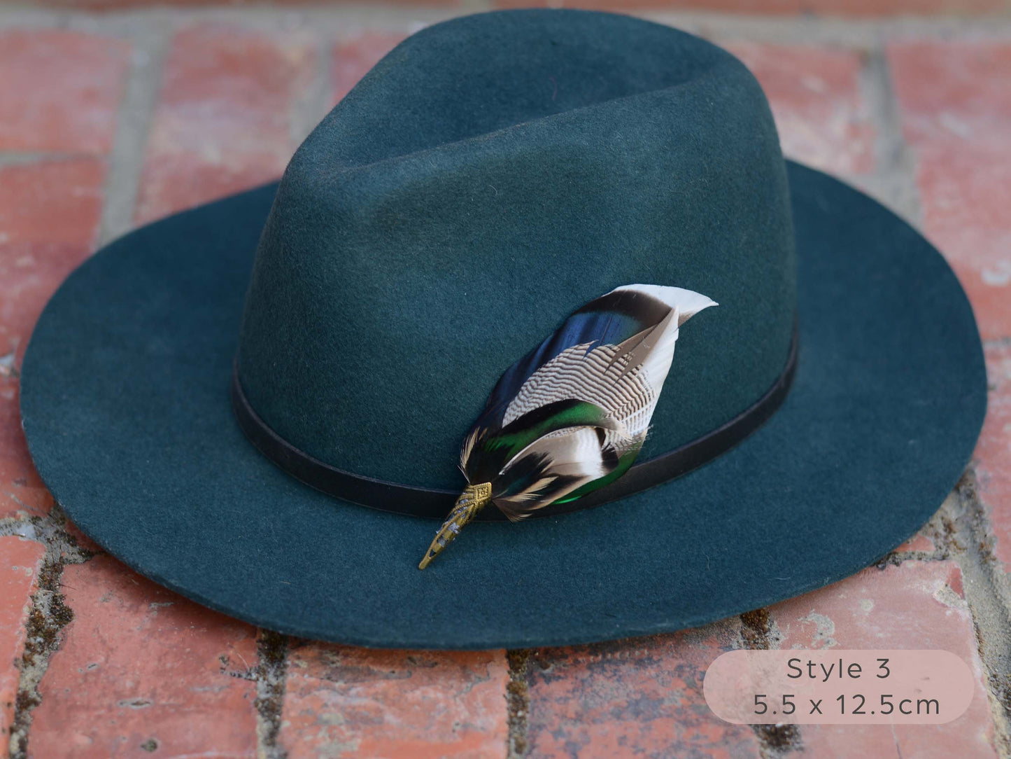 Duck feather hat pin with iridescent blue and green feathers on a dark teal hat with a thin black leather hat band on a red brick surface.
