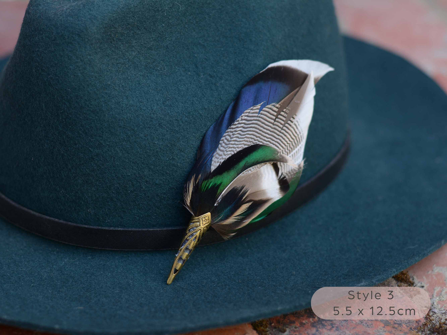 Close up of Duck feather hat pin with iridescent blue and green feathers on a dark teal hat with a thin black leather hat band on a red brick surface.