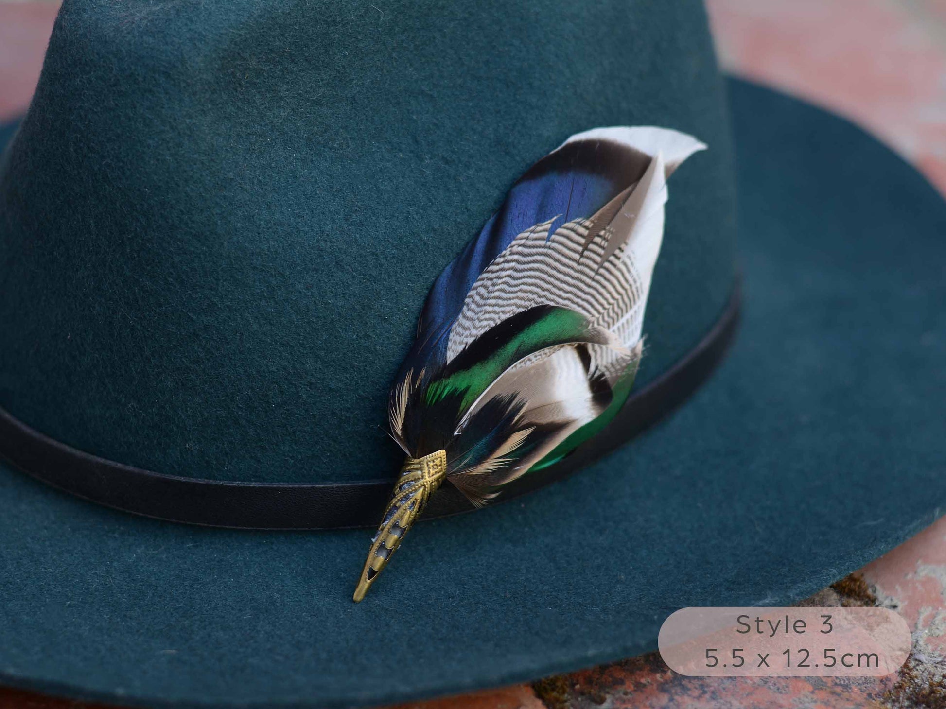 Close up of Duck feather hat pin with iridescent blue and green feathers on a dark teal hat with a thin black leather hat band on a red brick surface.