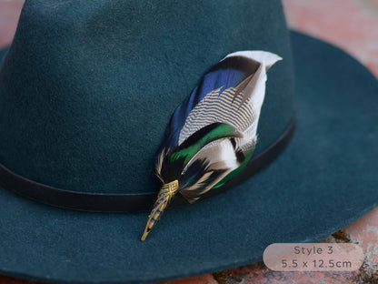 Close up of Duck feather hat pin with iridescent blue and green feathers on a dark teal hat with a thin black leather hat band on a red brick surface.