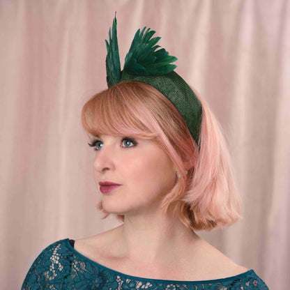 A woman with short pink hair wears a dark green fascinator headband decorated with two abstract bird "wings" crafted in green feathers, posed against a soft pink draped backdrop.