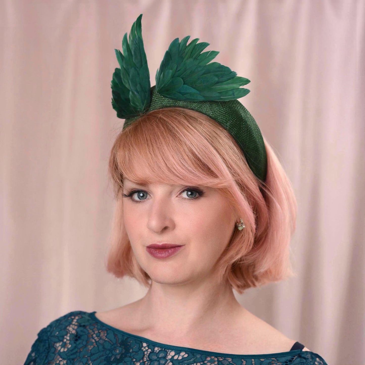 A woman with short pink hair wears a dark green fascinator headband decorated with two abstract bird "wings" crafted in green feathers, posed against a soft pink draped backdrop.