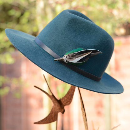 A teal felt wide-brim hat with a black band and a decorative feather duck feather hat pin, displayed outdoors on a metal stand against a blurred garden background.