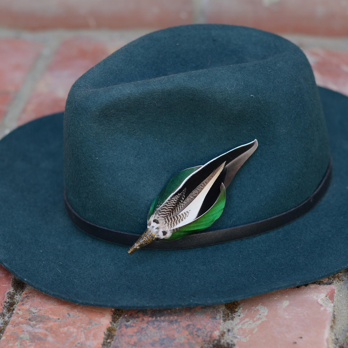 A dark teal felt fedora hat with a black band, decorated with a feather pin featuring green, black, and white feathers, resting on a red brick surface.