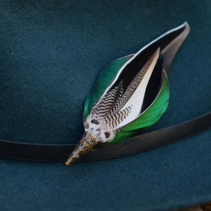 A decorative feather pin with iridescent green, black, white, and brown patterned feathers and a antique brass-toned cap, resting on a dark teal felt surface with a black band.