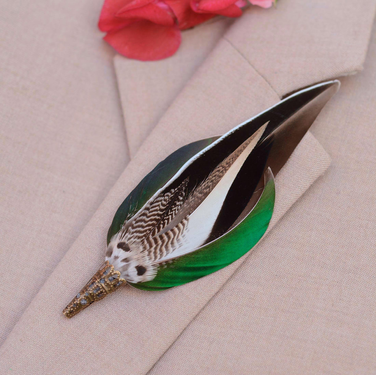 A decorative feather pin with a black-grey-and-white patterned feathers, and iridescent bright green wing feathers, resting on a beige textured fabric surface.