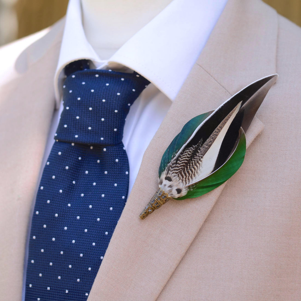 A close-up of a beige suit jacket on a mannequin or dress form, styled with a white dress shirt, a navy blue polka-dot tie, and a decorative feather lapel pin with green, grey, white and black duck feathers.