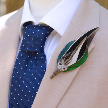 A close-up of a beige suit jacket on a mannequin or dress form, styled with a white dress shirt, a navy blue polka-dot tie, and a decorative feather lapel pin with green, grey, white and black duck feathers.