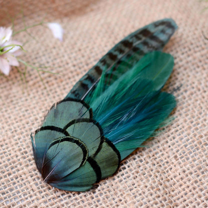 A decorative feather hair clip fascinator with iridescent teal, and green tones and black markings, arranged on a textured beige burlap surface with a small pale flower in the corner.