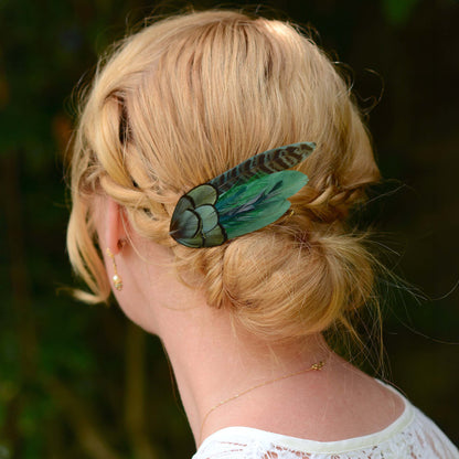 A blonde woman shown from the back wearing a green and black patterned feather hair accessory tucked into her low bun, with a dark blurred outdoor background.