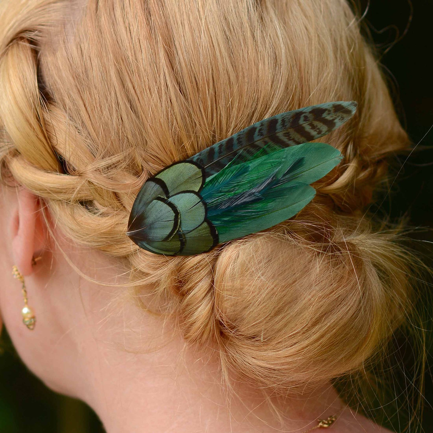 A close-up side view of a woman's hair styled in a low braid, featuring a decorative hair accessory with iridescent green and teal feathers attached to a black base. She is wearing a small gold dangle earring.