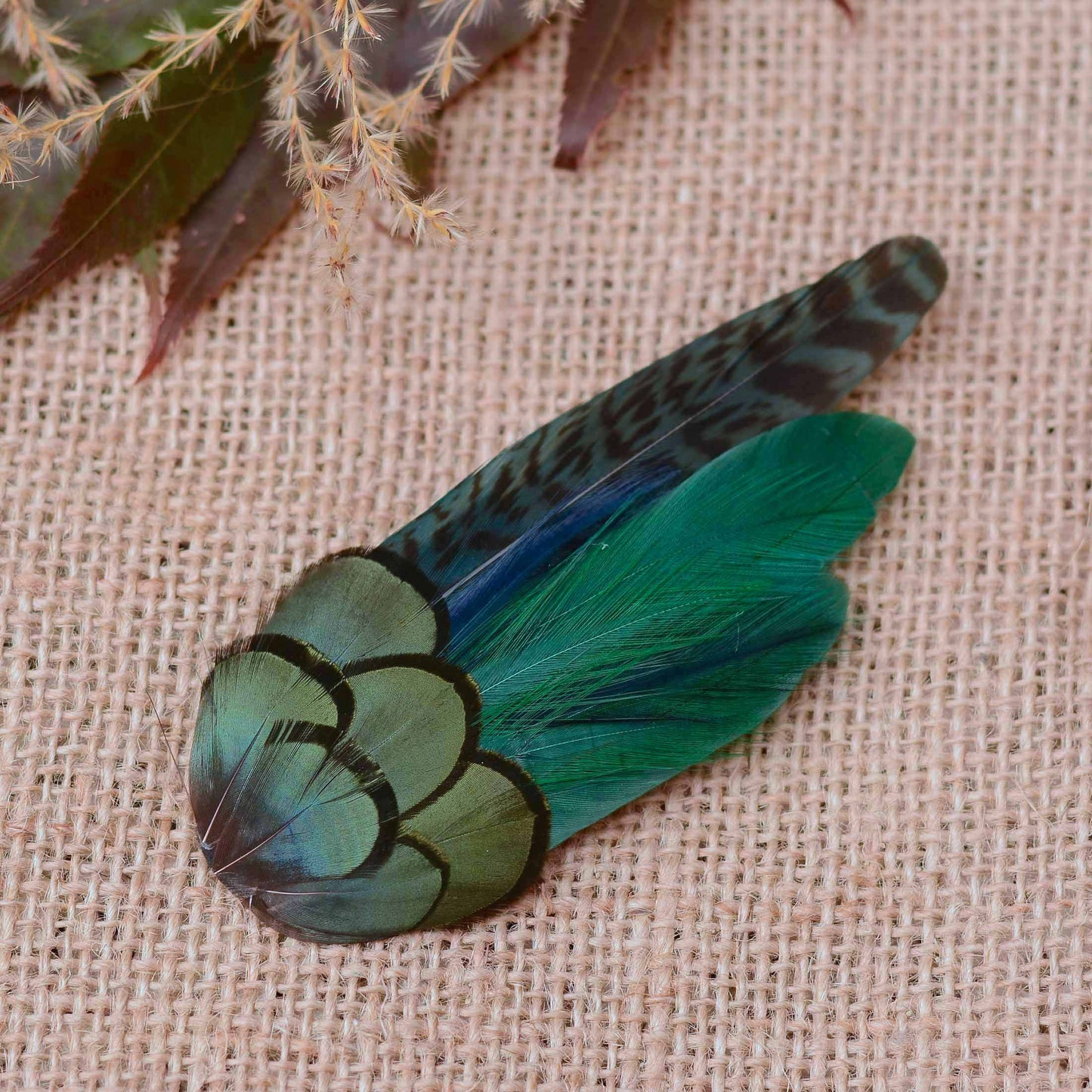 A decorative feather hair accessory with iridescent green and black feathers, placed on a beige burlap surface with dried plant stems in the corner.
