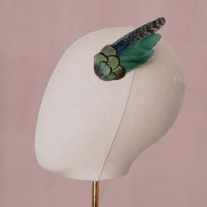 A decorative feather hair accessory with iridescent green and black feathers, displayed on a cream fabric mannequin head against a dusty pink background.