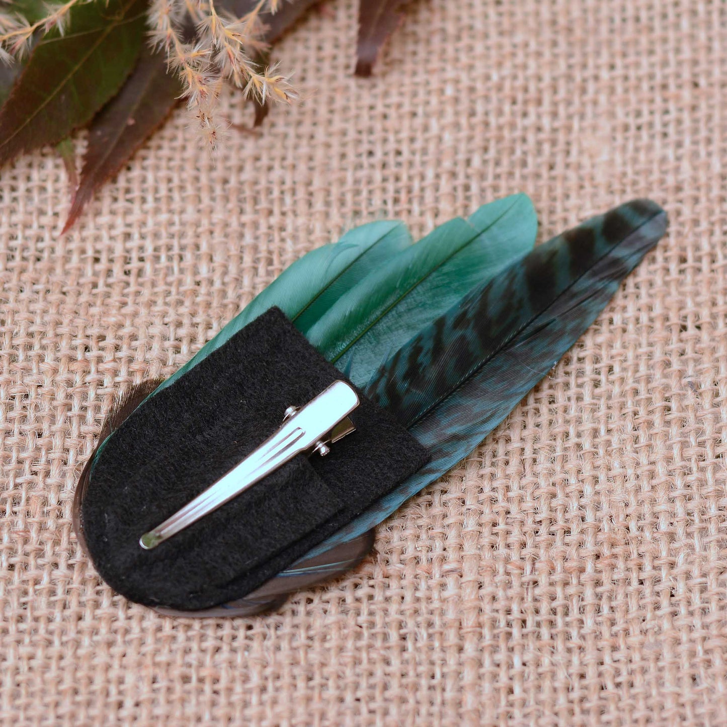 A hair clip decorated with green and black patterned feathers with a black felt backing and alligator metal hair clip resting on a textured beige burlap background.