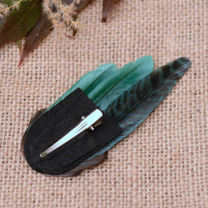 A hair clip decorated with green and black patterned feathers with a black felt backing and alligator metal hair clip resting on a textured beige burlap background.