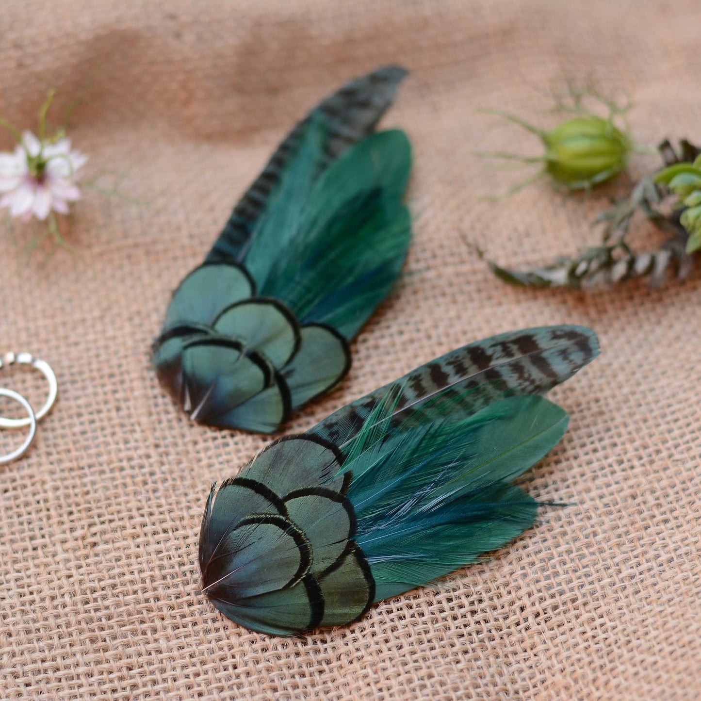 Two decorative feather hair clips in shades of teal, green, and black, displayed on a textured beige burlap fabric background with small flowers