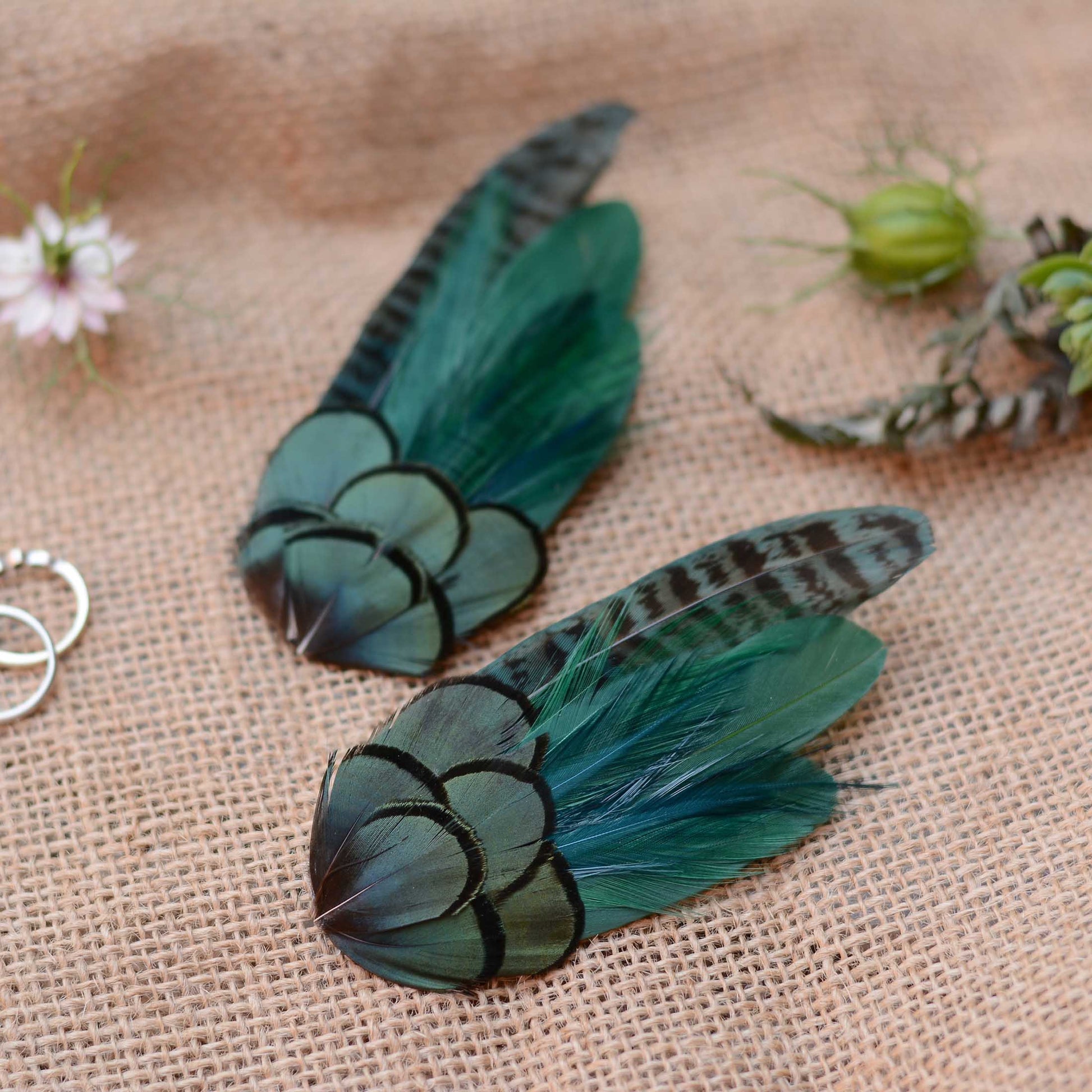 Two decorative feather hair clips in shades of teal, green, and black, displayed on a textured beige burlap fabric background with small flowers