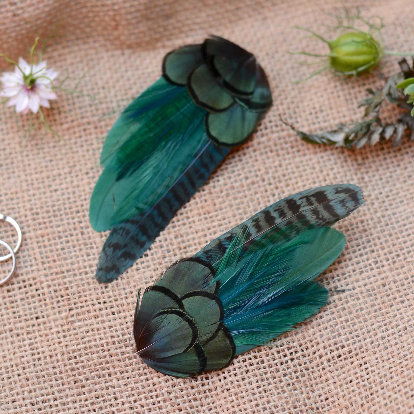 Two decorative feather hair clips in shades of teal, green, and black, displayed on a textured beige burlap fabric background with small flowers
