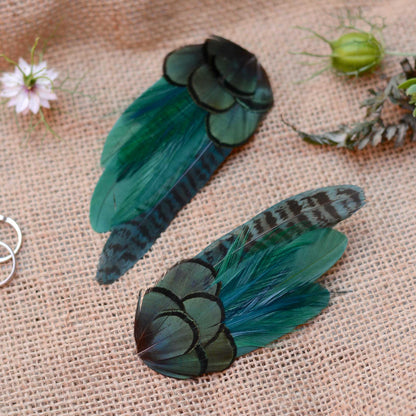 Two decorative feather hair clips in shades of teal, green, and black, displayed on a textured beige burlap fabric background with small flowers