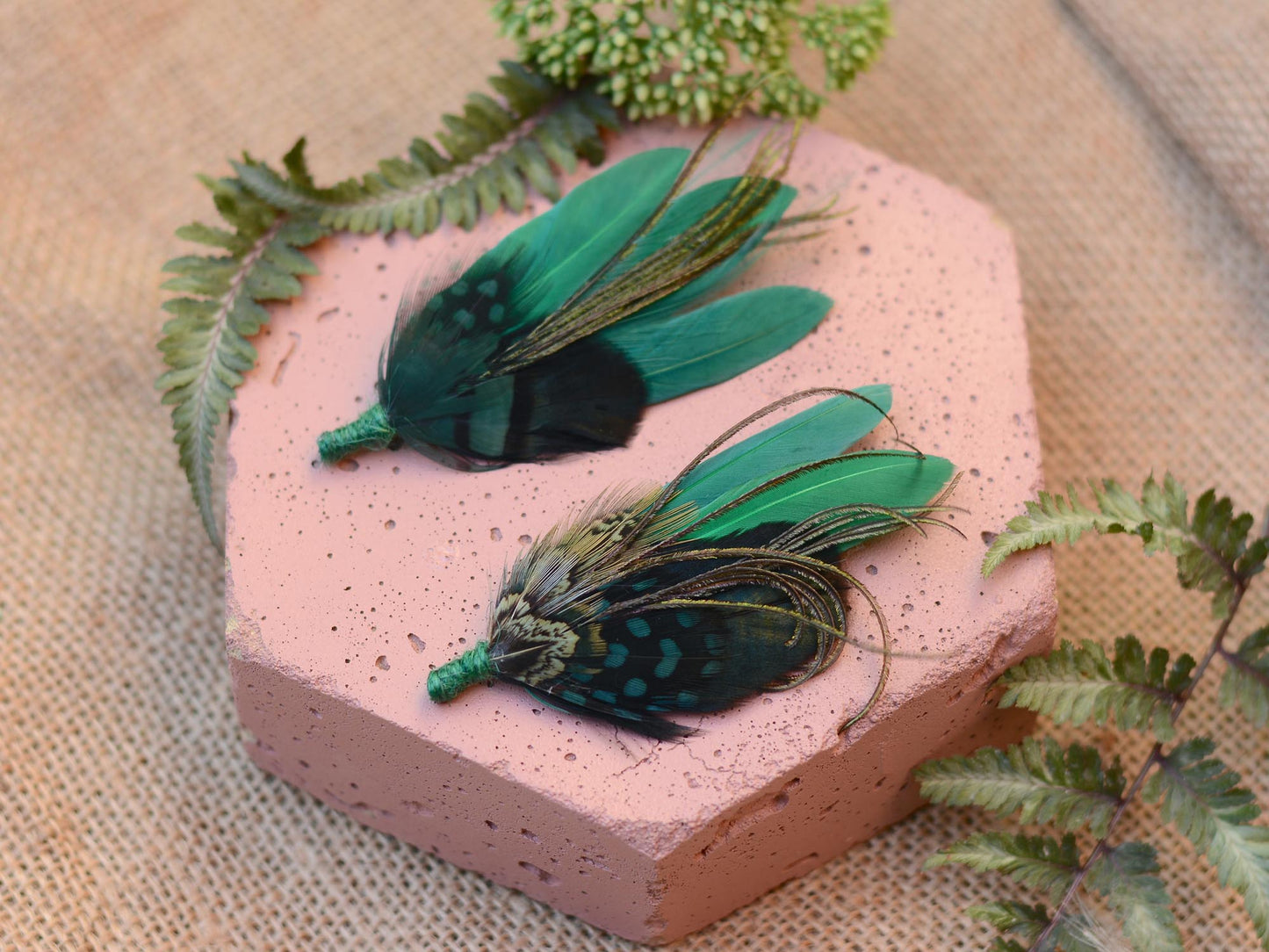Two green feathered boutonnieres on a pink stone and burlap background