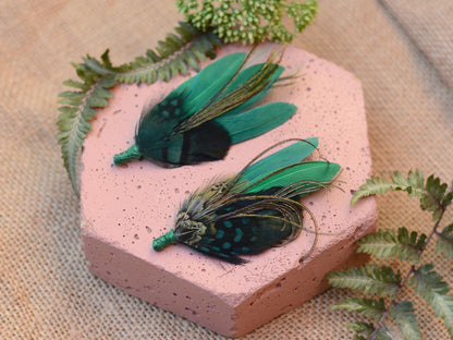 Two green feathered boutonnieres on a pink stone and burlap background
