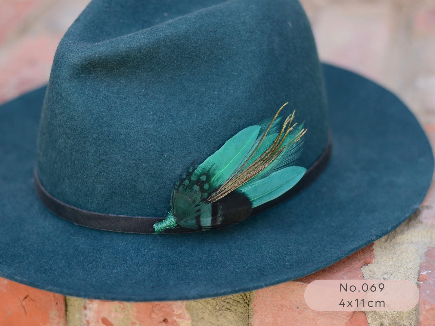 Teal fedora wool hat with a green pheasant, duck and peacock feather hat pin brooch on a  brick surface