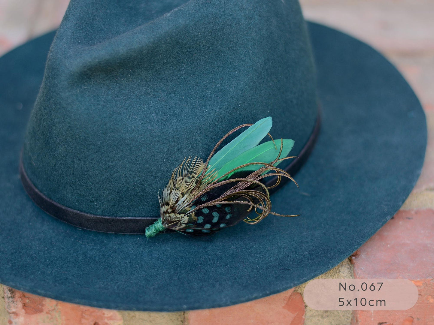 Teal fedora wool hat with a green pheasant, duck and peacock feather hat pin brooch on a  brick surface