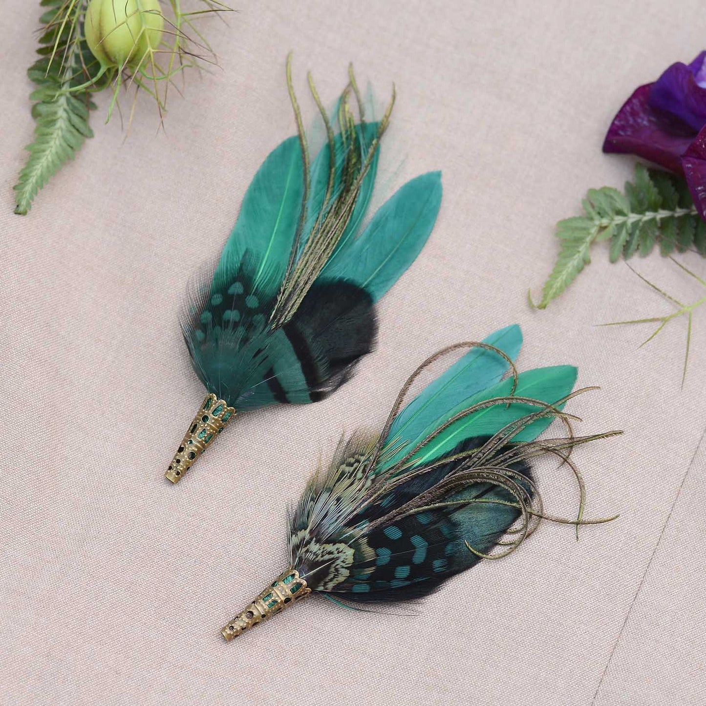 Two feathered brooches with green and black feathers on a light beige fabric surface with flowers and leaves.