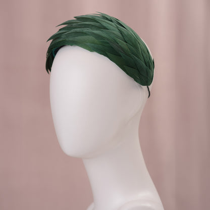 A dark green feather headpiece in a wide headband style. The feathers are cut to points and create texture and depth as they wrap around the head. The hat is fitted with a hat elastic. It is displayed on a white mannequin head against a soft pink background.