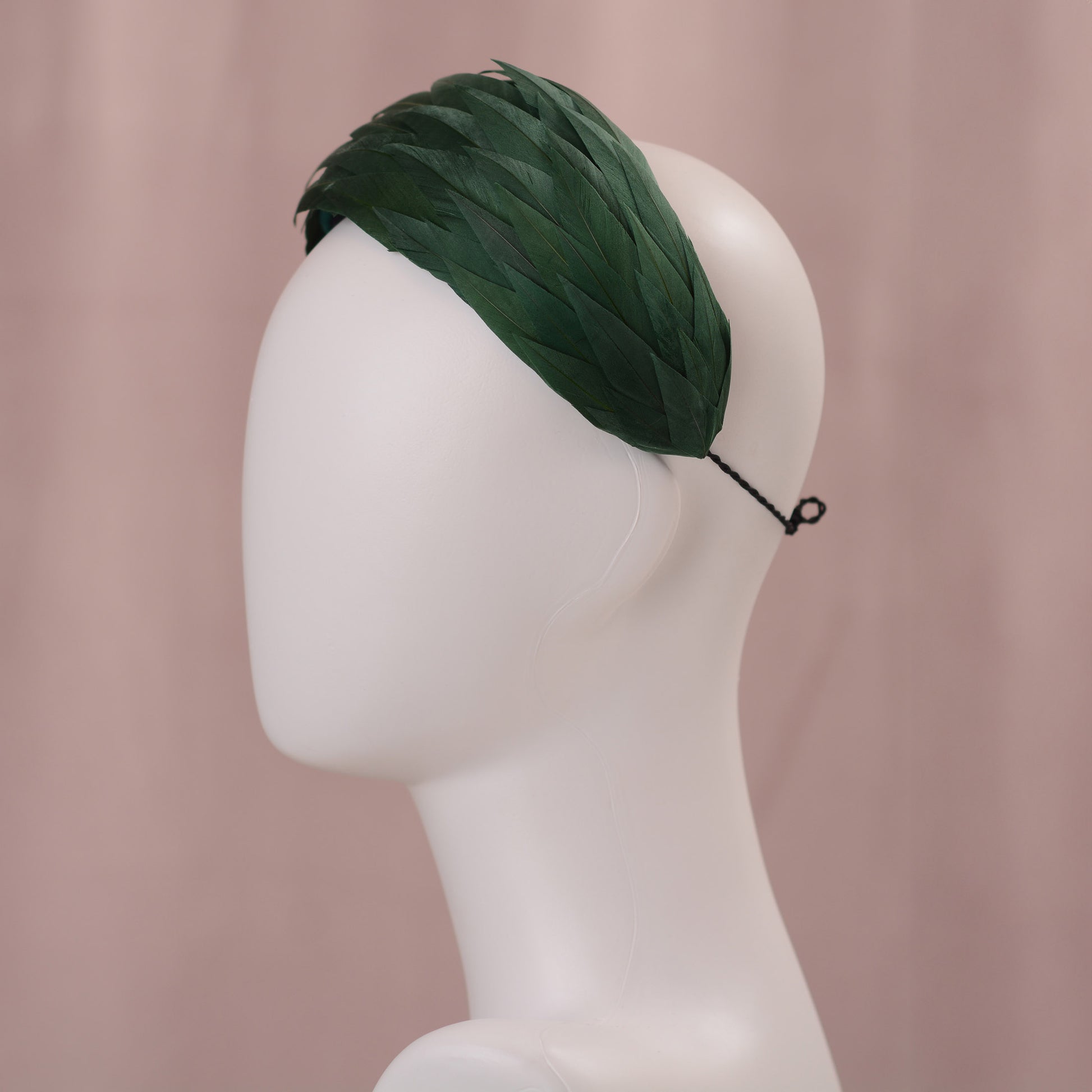 A dark green feather headpiece in a wide headband style. The feathers are cut to points and create texture and depth as they wrap around the head. The hat is fitted with a hat elastic. It is displayed on a white mannequin head against a soft pink background.